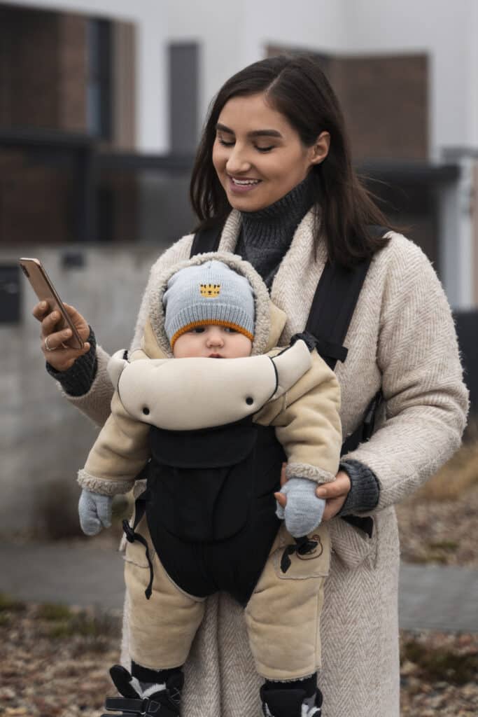 Woman walking holding a baby in a carrier using a smartphone.